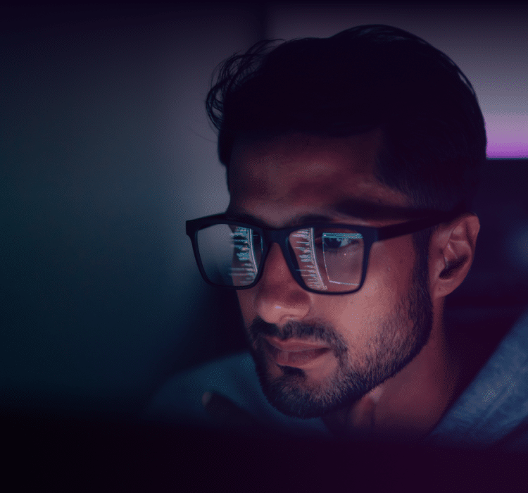 A man wearing glasses looks at a computer screen in a dimly lit room, the screen’s glow reflected in his glasses as he analyzes cybersecurity data for incident response.
