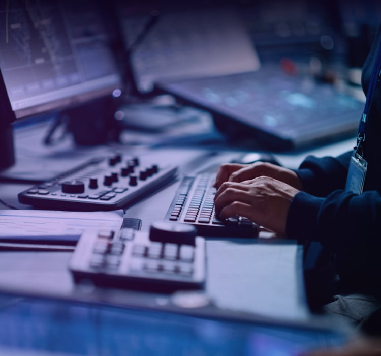 Person typing on a computer keyboard at a control desk with multiple monitors and equipment, suggesting an incident response or cybersecurity operations environment.