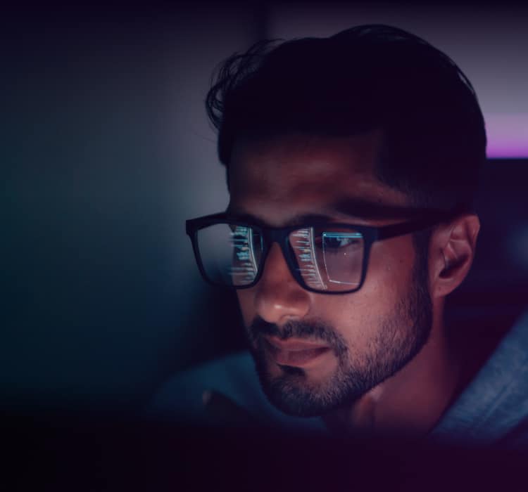 A man with glasses is looking intently at a computer screen in a dimly lit room, threat intelligence data and code reflected in his lenses as he analyzes enterprise security.